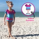 Young girl at beach wearing Fuchsia Turquoise Ombre short sleeve swimsuit by FASTEN. Rash guard swimsuit. Fits toddler girls and young girls, sizes 2T-10. Features patented design that opens at the waist, making bathroom breaks and baby diaper changes faster and easier. UPF 50 sun protection built right in! Ombre varies slightly in each swimsuit. 